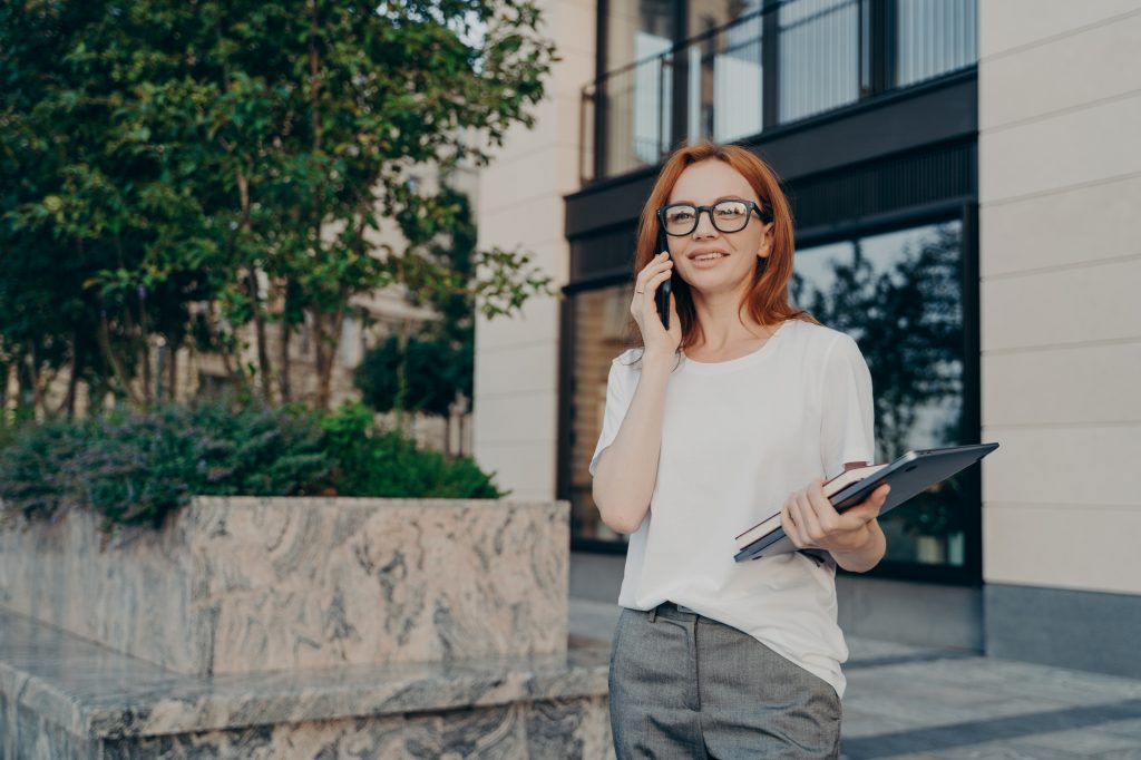 Young smiling european businesswoman calling on smartphone while walking in city with laptop in hand